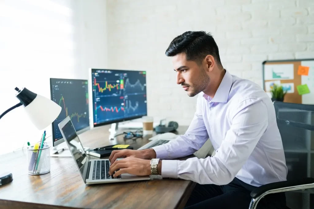 Homem de camisa social trabalhando em home office, digitando em notebook à frente de três telas mostrando gráficos de velas e indicadores financeiros