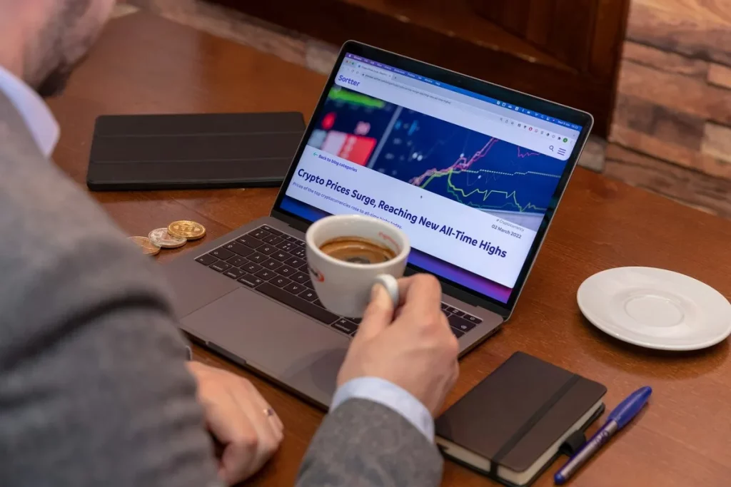 Pessoa analisando gráfico de criptomoedas em notebook com notícia sobre alta histórica, ao lado de moedas físicas de Bitcoin, representando estudo de risco e retorno com base no Índice de Sharpe.