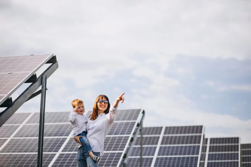 Mulher segurando criança pequena sorri e aponta ao lado de painéis solares.