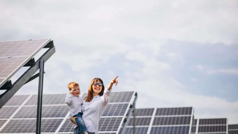 Mulher segurando criança pequena sorri e aponta ao lado de painéis solares.