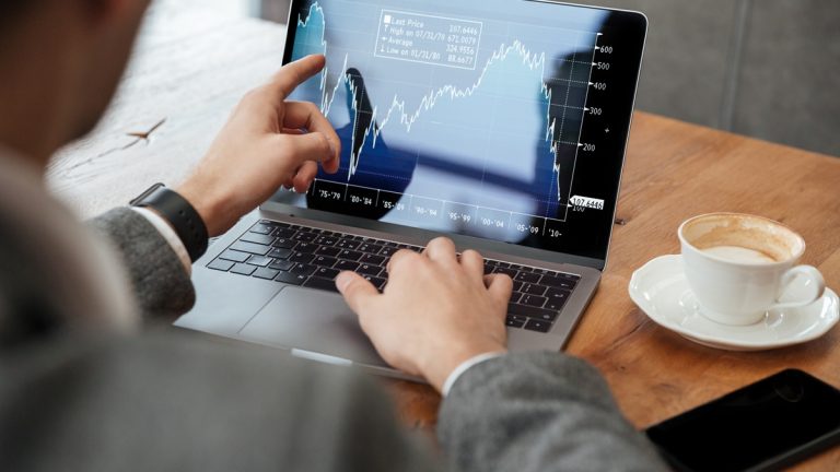 Imagem de uma pessoa analisando gráficos financeiros no laptop enquanto aponta para a tela. Ao lado, há uma xícara de café e um celular.