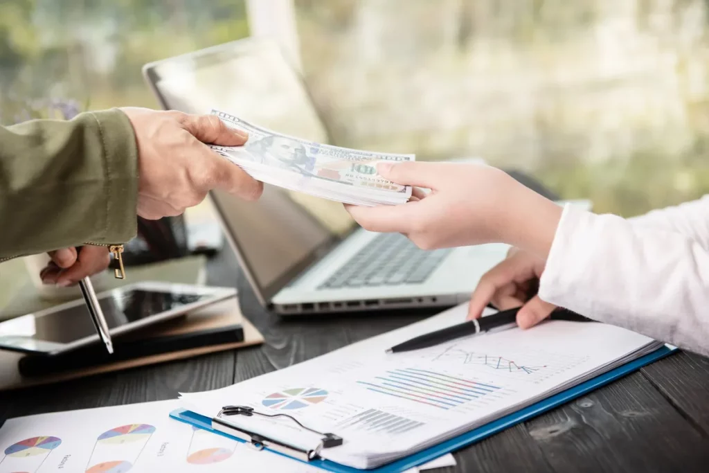 Pessoa entregando maço de dólares para outra em mesa com gráficos financeiros e notebook.