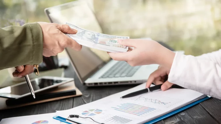 Pessoa entregando maço de dólares para outra em mesa com gráficos financeiros e notebook.