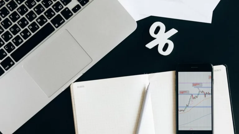 Mesa de trabalho com laptop, smartphone exibindo gráficos de investimento, caderno, lápis e um símbolo de porcentagem, representando o planejamento financeiro para CDB para investir.