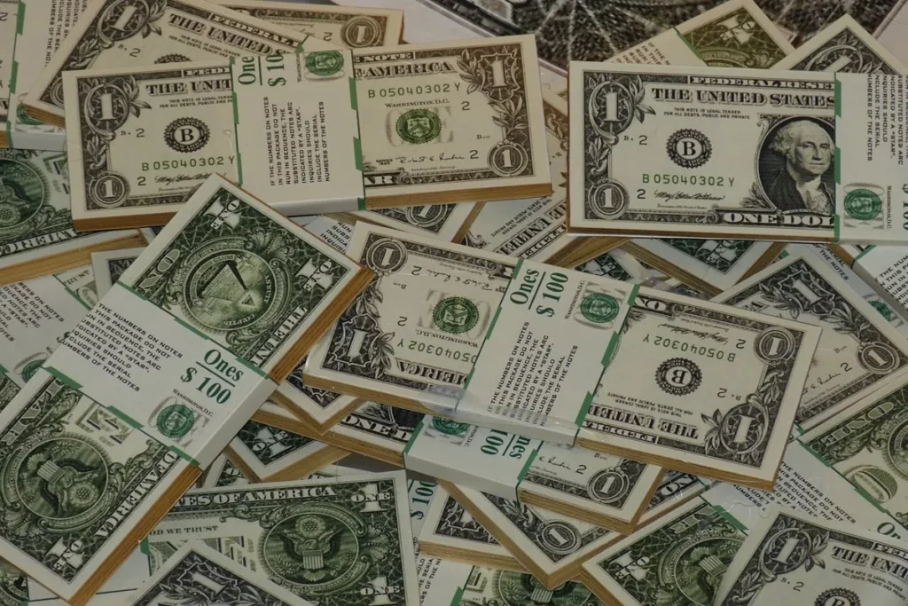 Pilhas e notas soltas de dólares americanos, com destaque para cédulas de $1 e maços de $100, simbolizando como investir em dólares.