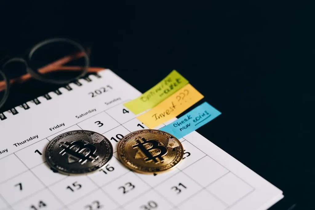 Duas moedas de Bitcoin (uma prateada e uma dourada) sobre um calendário de 2021, com notas adesivas coloridas com lembretes como "Oportunidade de mercado", "Investir agora" e "Verificar novas moedas". Um par de óculos está no canto superior esquerdo. Esta imagem representa o planejamento financeiro e oportunidades para investir para iniciantes em criptomoedas.