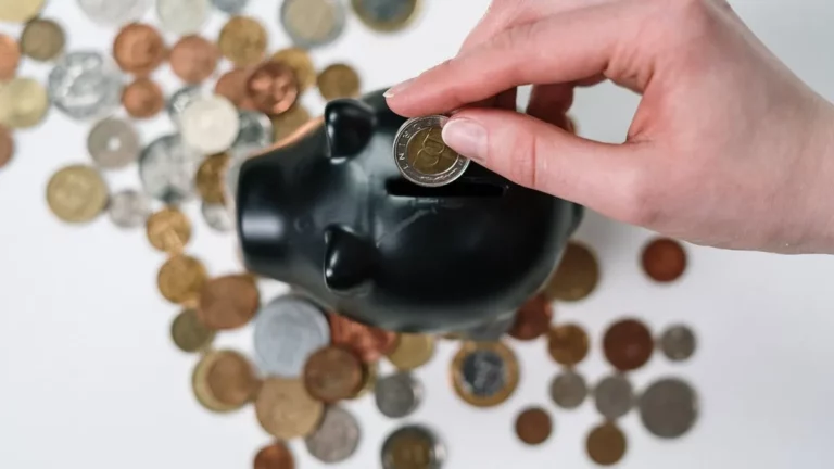 Mão inserindo uma moeda de dois euros em um cofrinho preto em formato de porco. Várias outras moedas de diferentes países e valores estão espalhadas sobre uma superfície branca ao redor do cofrinho. Esta imagem conceitua a poupança como um passo fundamental para investir para iniciantes.