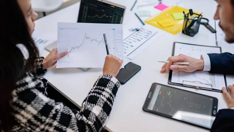 Duas pessoas em reunião de negócios analisando gráficos financeiros e tablets, discutindo qual é a melhor corretora para investir.