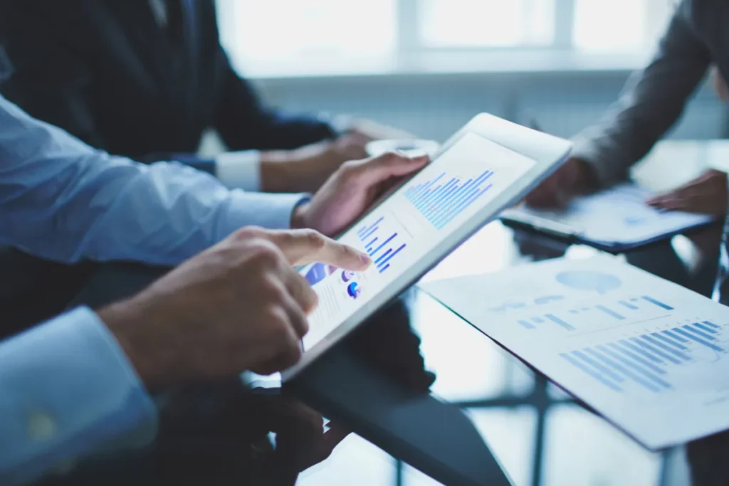 Profissional analisando gráficos e dados financeiros em tablet durante reunião de negócios, buscando qual é a melhor forma de investir dinheiro.