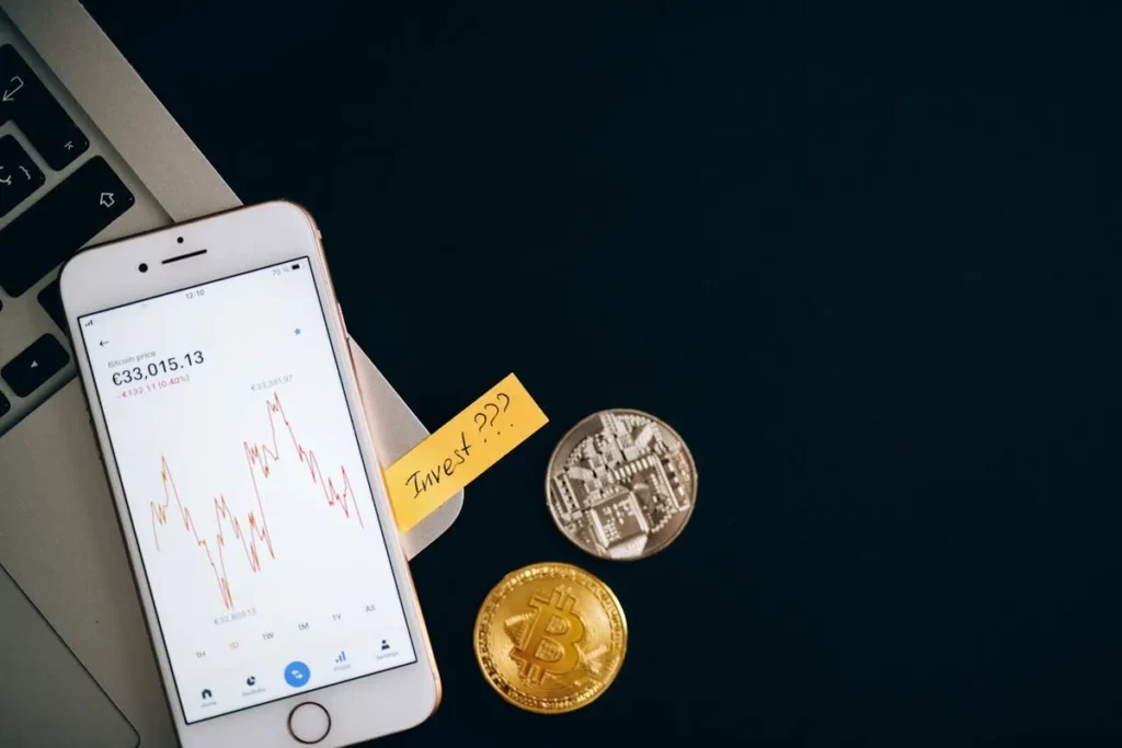 Um smartphone exibindo um gráfico de queda de preço do Bitcoin (€33,015.13 e -1.10%) ao lado de moedas físicas de criptomoeda (uma dourada de Bitcoin e uma prateada), com um post-it amarelo escrito "Invest???" preso à tela, representando a volatilidade do mercado de criptomoedas e a necessidade de "stop loss".