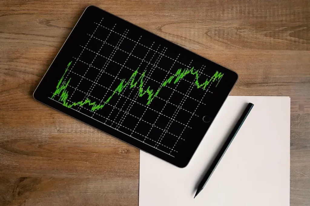 Um tablet exibindo um gráfico de linha verde em uma grade preta, simulando dados de mercado financeiro, posicionado em uma mesa de madeira ao lado de uma folha em branco e um lápis preto, ilustrando o planejamento de investimentos e o conceito de "stop loss".