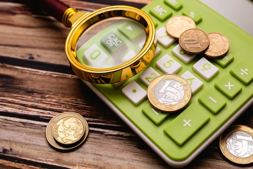 Uma lupa dourada sobre uma calculadora verde, com moedas de real espalhadas, simbolizando o planejamento e a análise financeira para investimentos.