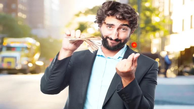 Homem de terno, com barba, sorrindo e segurando um maço de dinheiro, fazendo um gesto de "venha cá", com fundo de rua de cidade.