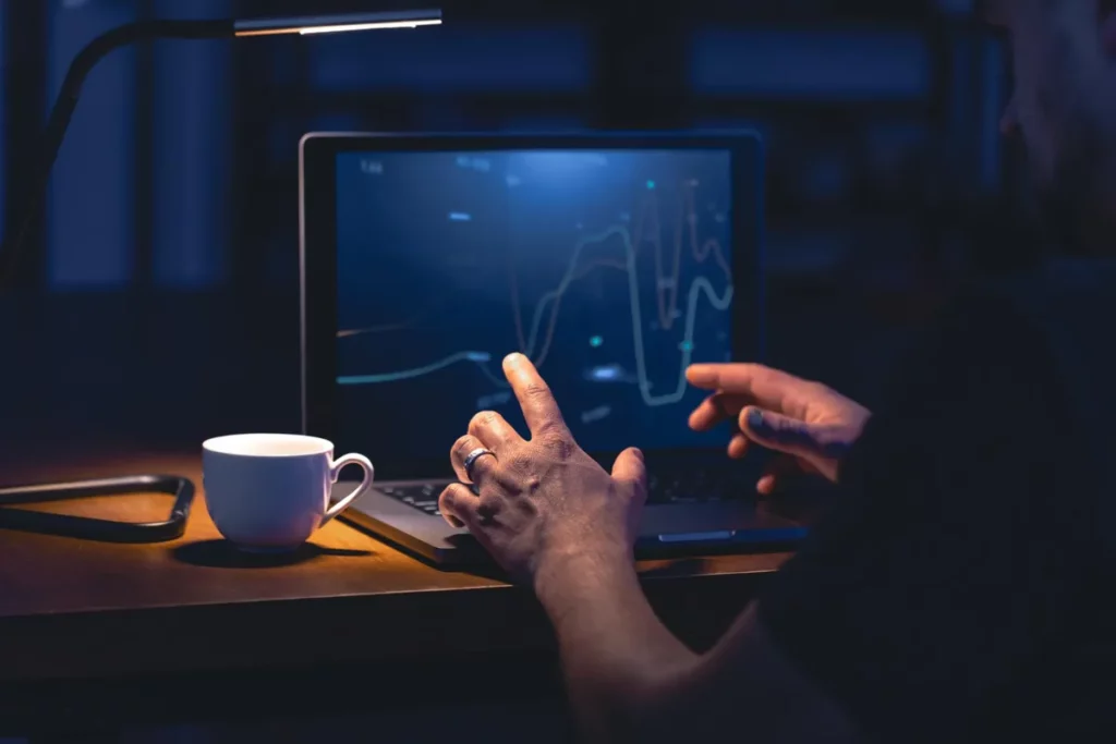 Pessoa de mãos sobre um notebook, com um anel no dedo, trabalhando no escuro. A tela do computador exibe um gráfico financeiro com linhas de tendência em cores claras, indicando uma análise de mercado ou sessão de trading. Uma luminária e uma xícara de café estão ao lado, sugerindo um trabalho noturno.