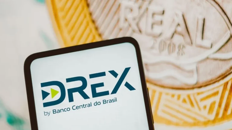 Um close-up de um smartphone exibindo o logo do DREX ("DREX by Banco Central do Brasil") na tela. O smartphone está posicionado em frente a uma nota de dinheiro e uma moeda de Real brasileiro desfocadas. A imagem destaca a interface digital da nova moeda DREX e sua conexão com o dinheiro físico.