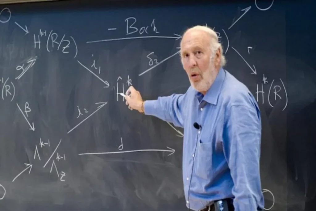 Retrato de Jim Simons, vestindo uma camisa azul, gesticulando com giz em uma lousa coberta por diagramas de matemática avançada e equações.