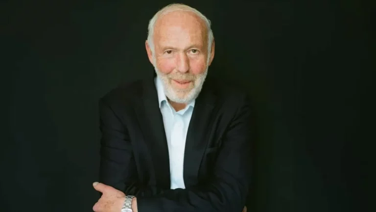 Retrato de Jim Simons, sorrindo, vestindo um blazer escuro e camisa clara, em frente a um fundo preto.