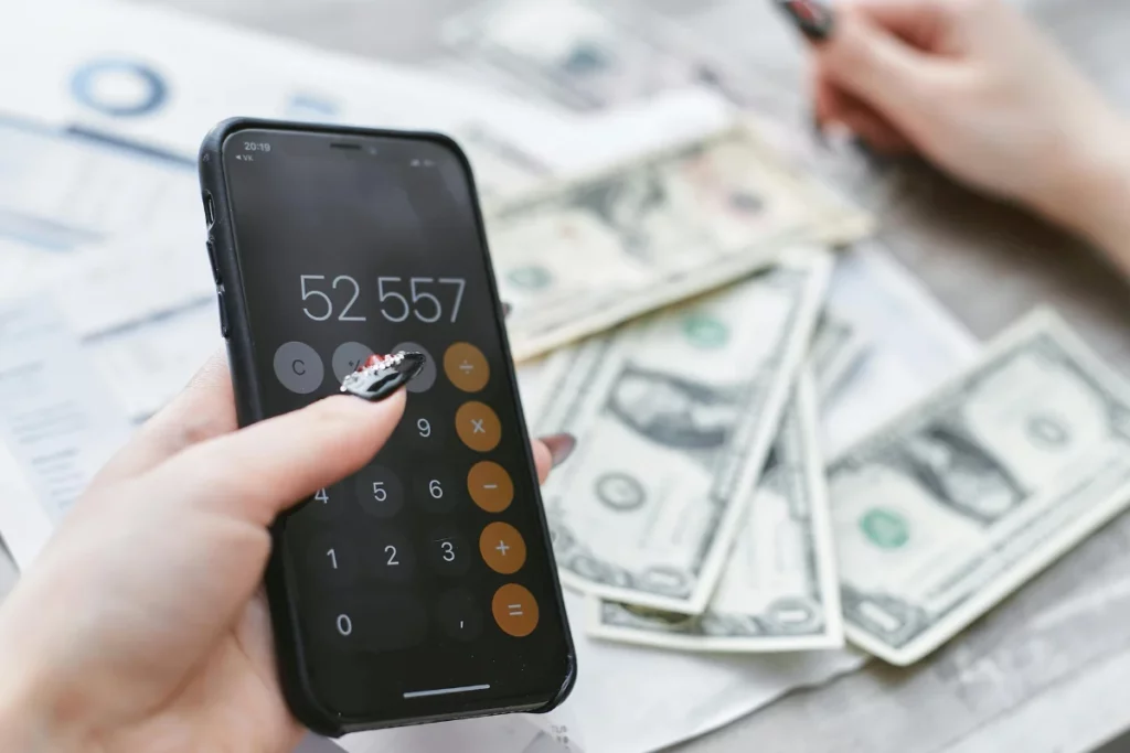 Close-up de uma mão segurando um smartphone preto exibindo a calculadora com o número 52557. Em segundo plano, há notas de dólar (US$1) e documentos em uma mesa, representando gestão financeira e contabilidade.