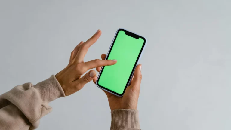 Close-up de mãos segurando um smartphone moderno com bordas brancas e tela verde brilhante (chroma key), prontas para substituição de conteúdo. Um dedo indicador toca o centro da tela, sugerindo interação ou toque.