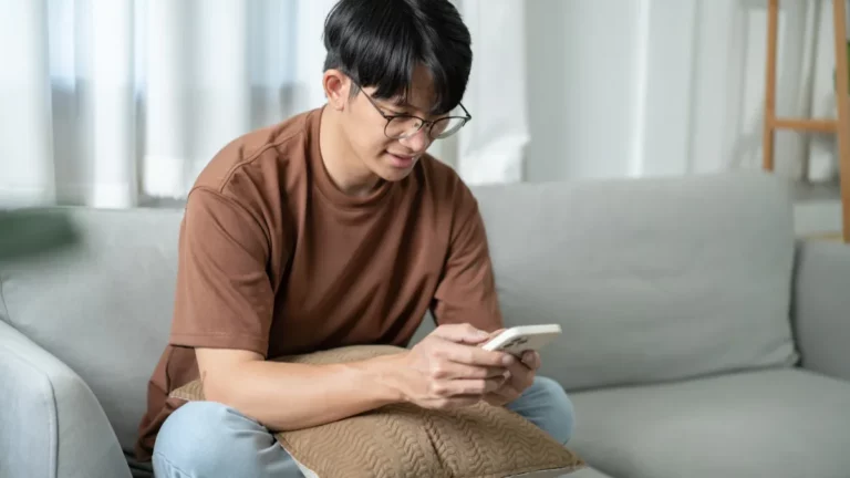 Homem sentado no sofá olhando para o celular enquanto navega ou faz uma compra online.