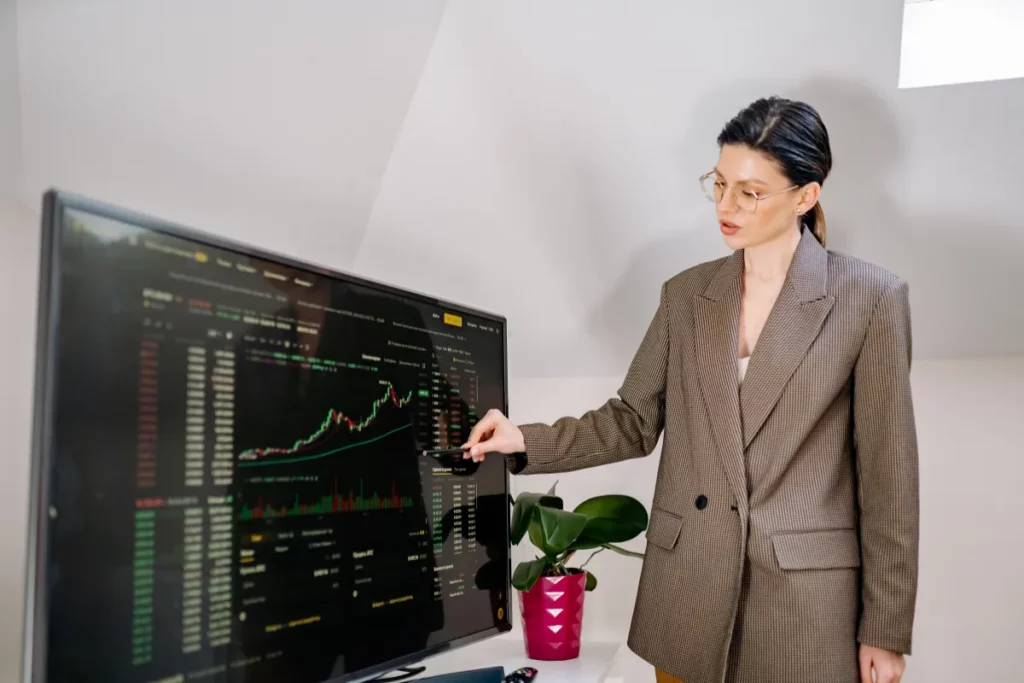 Mulher apontando gráfico de ações em tela grande com dados financeiros exibidos